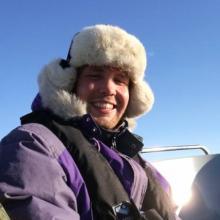 Steffen Ringsø Nielsen - pale skinned man wearing a fur-lined hat and a purple puffy jacket smiles at the camera outside with blue sky above