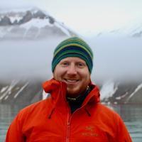 Man in striped wool hat and orange rain jacket stands in front of fjord with mountains in fog