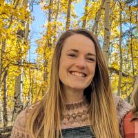 Mariama (woman with fair skin, freckles, and light brown hair, wearing a pink and brown wool sweater and denim overalls) smiles at the camera in front of an aspen grove