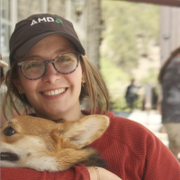 Robyn Marowitz (woman with fair skin, glasses and brown hair pulled back into a ponytail, wearing a hat and a red top, and holding a corgi) smiles at the camera
