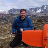 Frederick Hass (man with pale skin, brown hair and a brown beard) smiles at the camera in front of Greenlandic mountains, holding an orange drone in the foreground